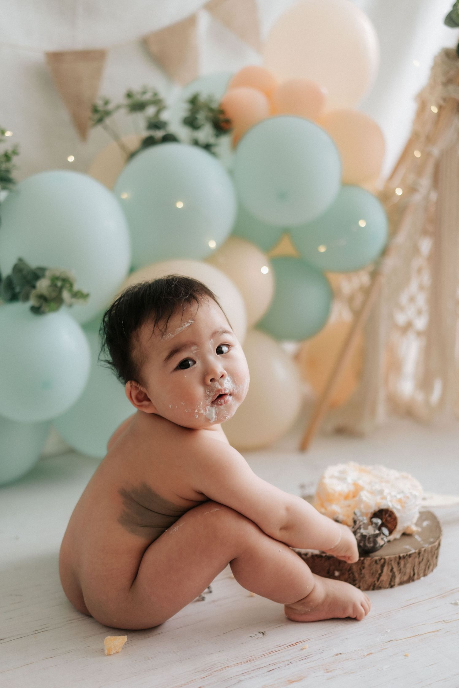 Baby cake smash photography session with mint green and peach balloons against white backdrop and rustic wooden decor.
