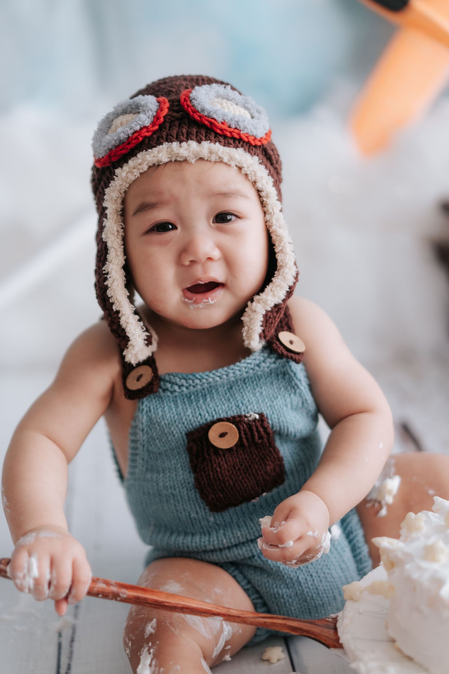 Baby wearing blue knit vest and aviator style winter hat plays with baking ingredients and wooden spoon.