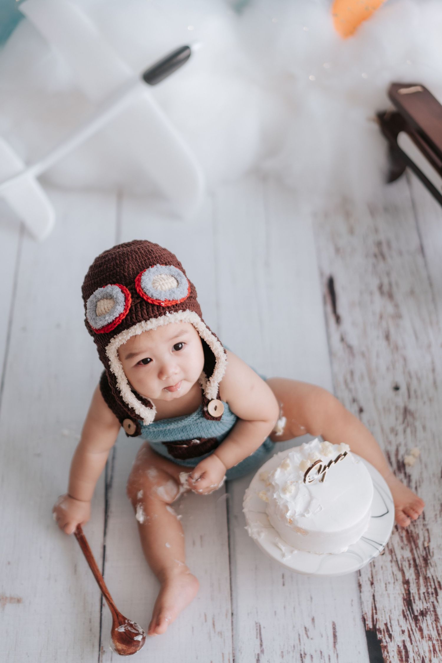 A baby wearing a knitted aviator hat plays with white frosting on a rustic white wooden surface.