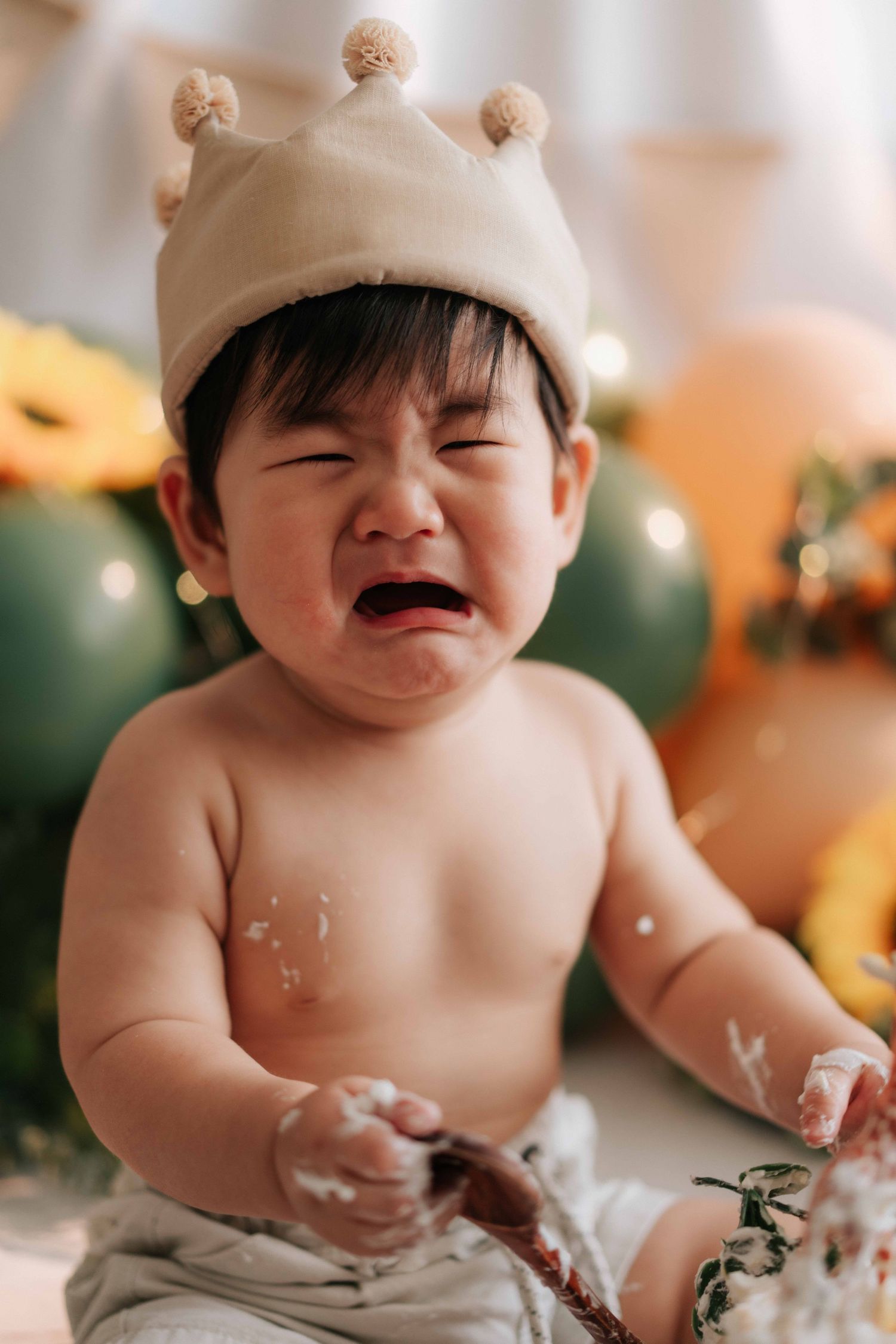 A happy baby wearing a beige hat plays and laughs while making a mess with cake during a smash cake photo session.