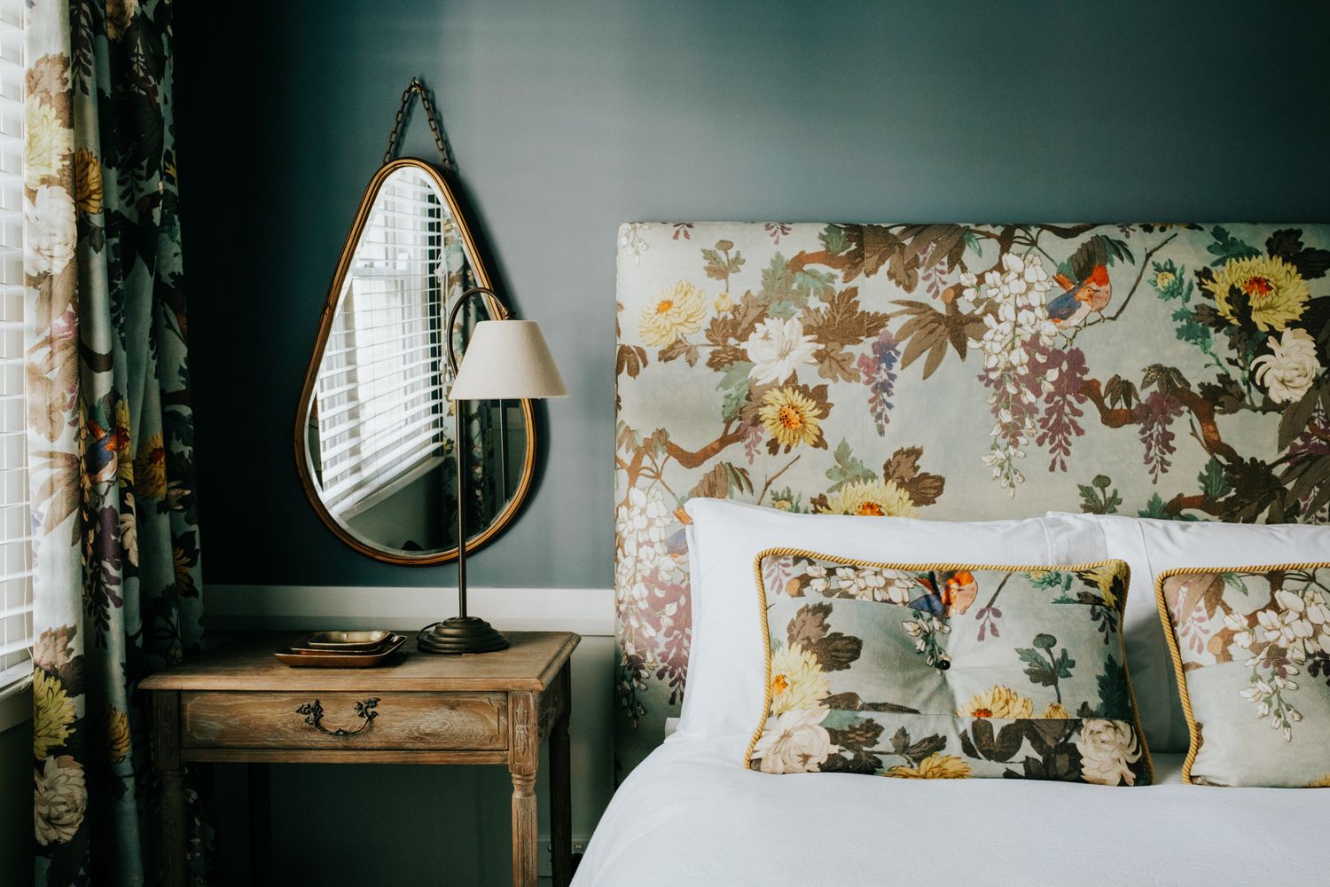 Floral wallpaper and vintage decor create a cozy bedroom interior with natural light streaming through window blinds.