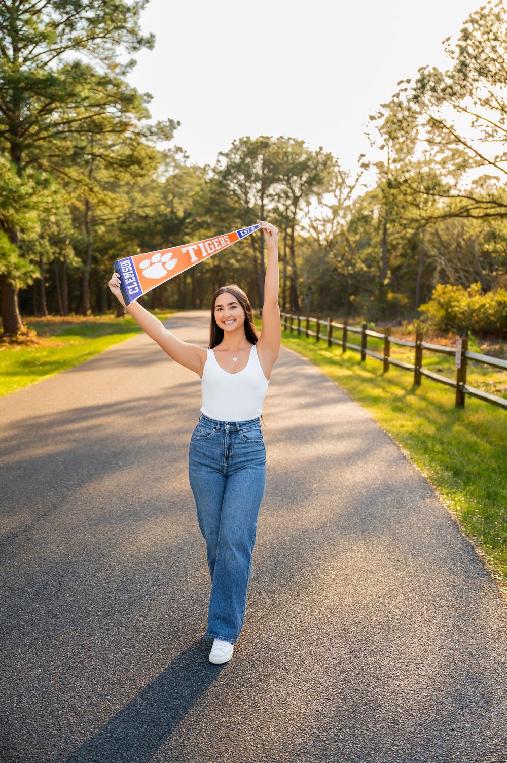 A person in jeans and white tank top poses playfully on a tree-lined path while holding a Clemson Tigers pennant.