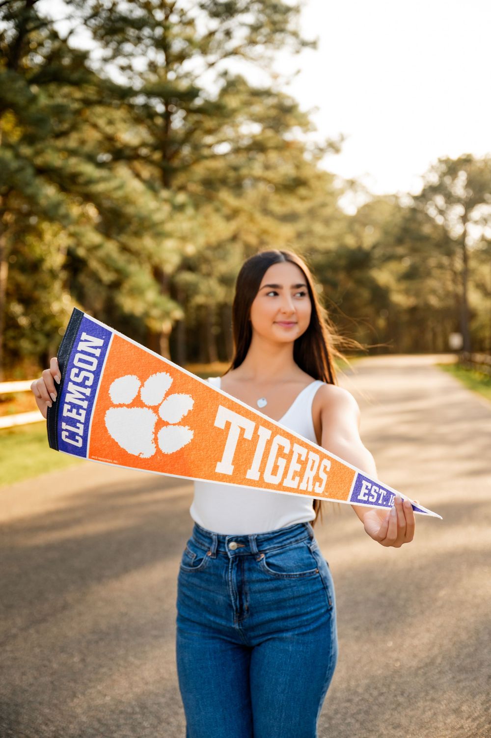 Person holding orange and white Clemson Tigers pennant on a sunlit path surrounded by trees.