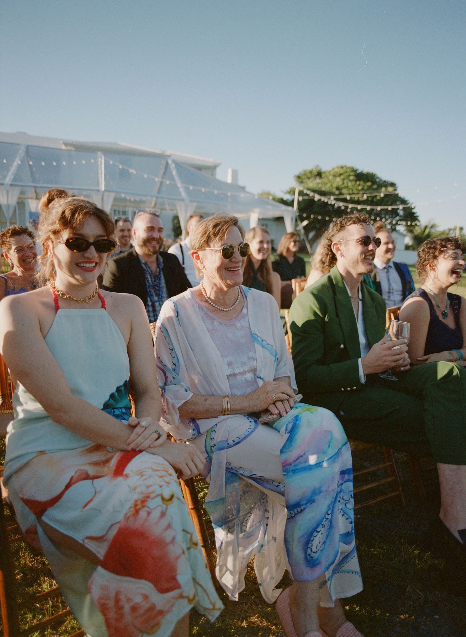 Wedding guests sit outdoors in white chairs during a sunny ceremony with ocean views in the background.