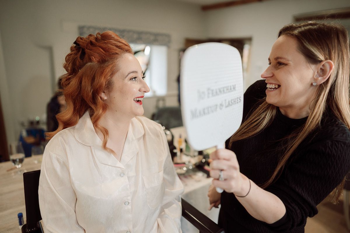 A hairstylist works on styling a bride's red hair during wedding preparations in a bright salon setting.