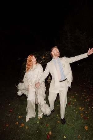 A couple in white formal attire dances playfully on a grass lawn at night with fallen autumn leaves scattered around.