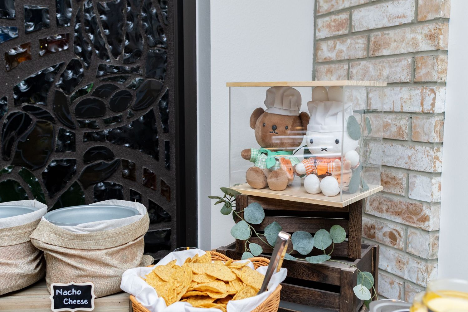 A rustic party spread features a charcuterie board, snacks, and drinks arranged on a wooden table against a stone wall.