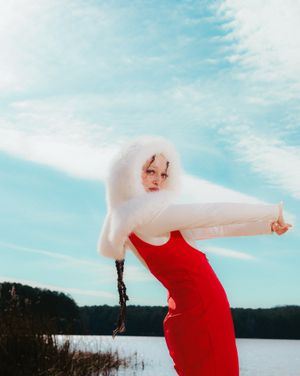 A figure in a flowing red dress dances gracefully against a serene lakeside backdrop with blue sky and clouds.