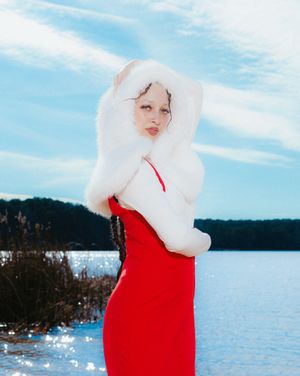 A figure in a bright red dress and white headpiece stands against a serene lake and blue sky backdrop.