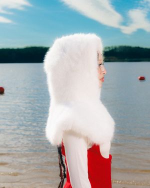 A figure in a red swimsuit and dramatic white fur coat poses dramatically by a lake on a sunny day.