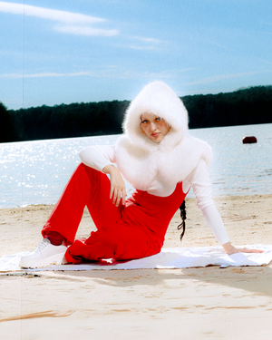 A figure in a red dress and white fur coat poses dramatically on a sandy beach near a lake.