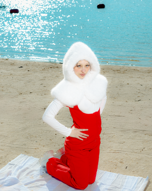 A figure in a red dress and white wig sits gracefully on a white blanket at the beach with turquoise water in the background.