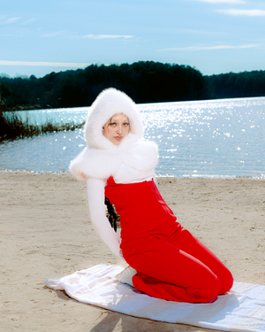 A figure in a bright red jumpsuit and white hair lounges on a white blanket at a sandy beach with water in the background.
