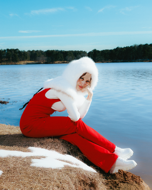 A figure in red clothing does a backbend movement on a sandy lakeshore with blue water and trees in the background.