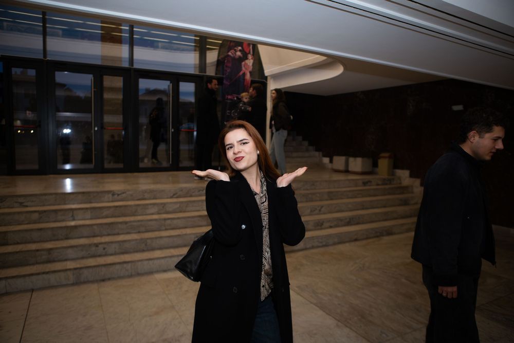 Young woman in black coat gesturing with hands in theater lobby.