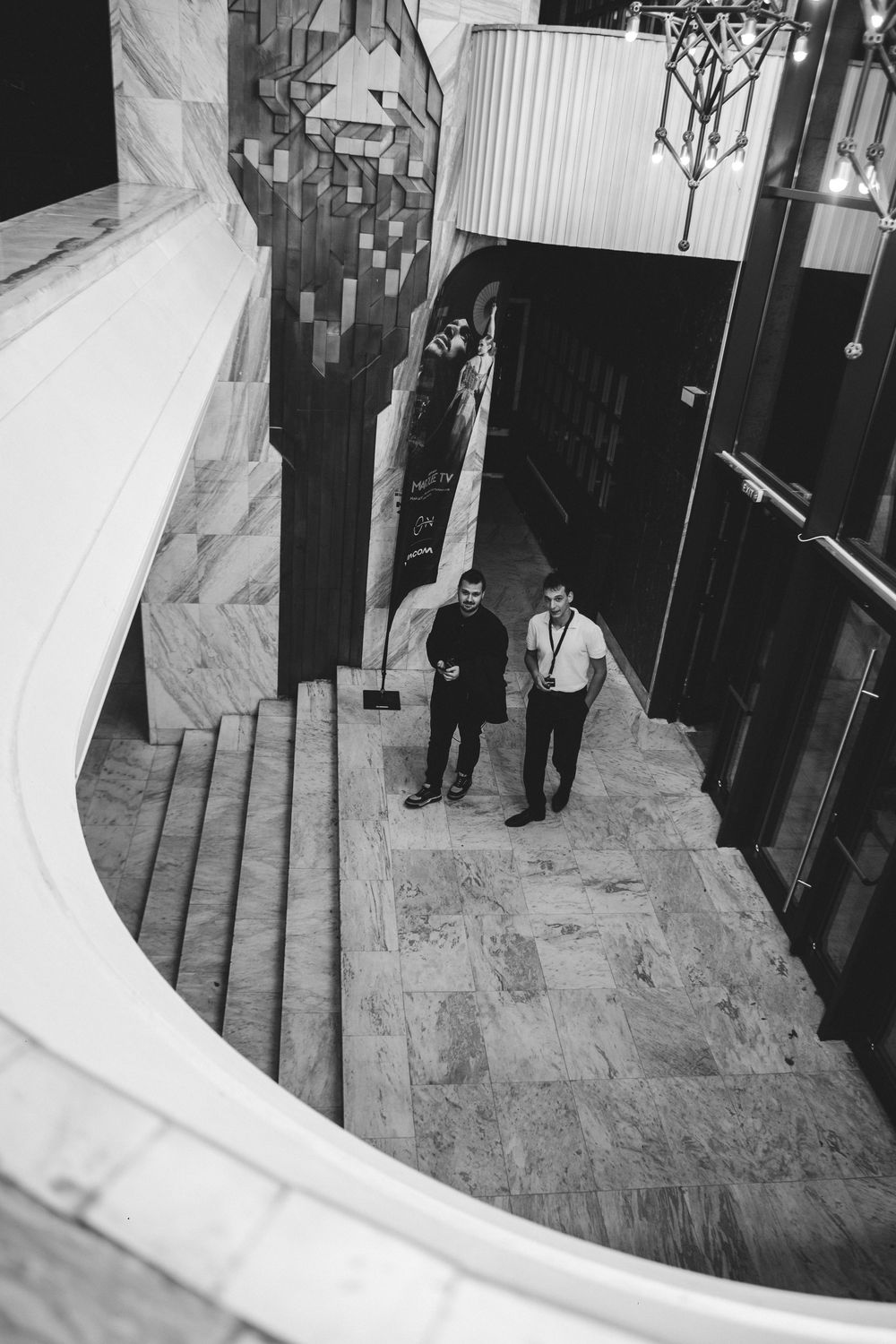 Black and white aerial view of two men standing near grand staircase in elegant lobby.