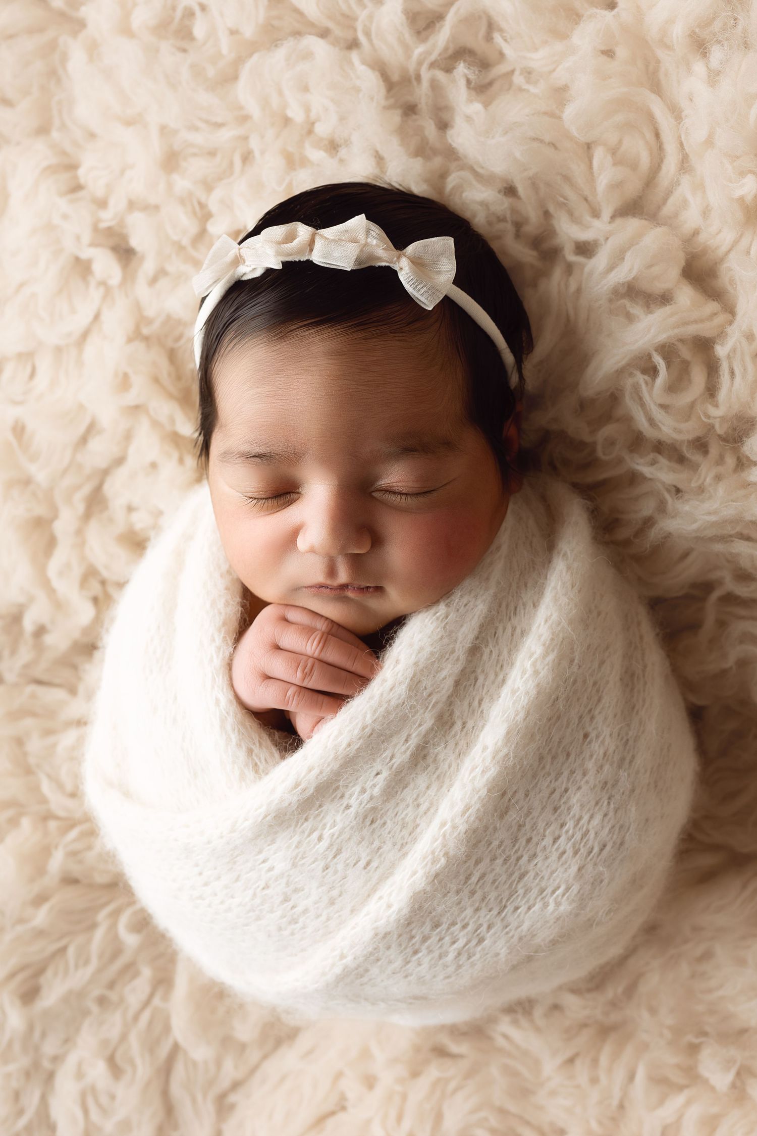 Sleeping newborn wrapped in soft white knit blanket with bow headband lying on cream textured backdrop.