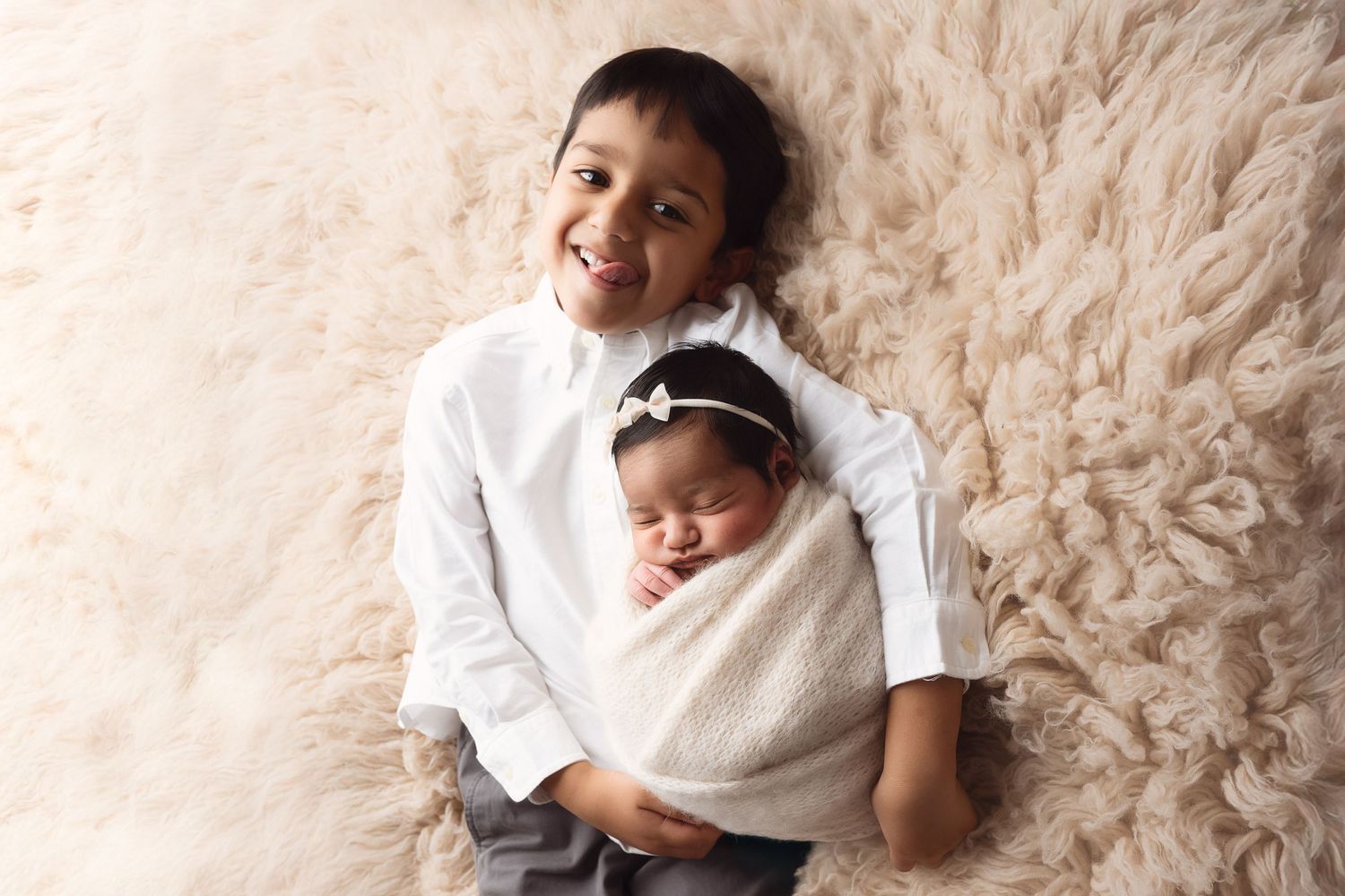 A sibling duo poses on a fluffy cream rug during a newborn photo session.