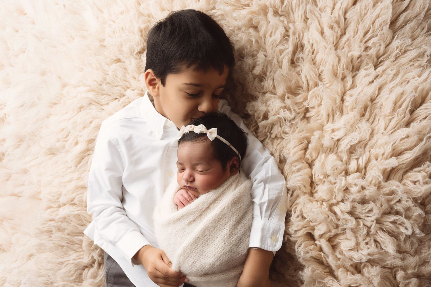 A baby wearing a white outfit lies on a plush beige carpet while looking up with a sweet expression.