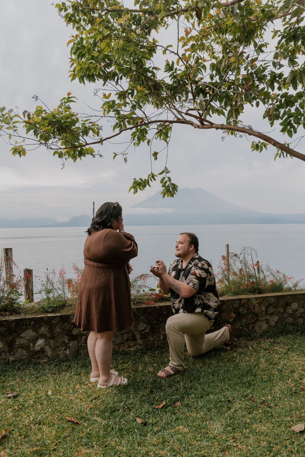 Person kneeling to propose to partner outdoors by a scenic lake with mountains in the background.