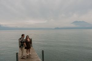 Couple walking hand in hand on a wooden dock overlooking a serene lake with misty mountains in the background.