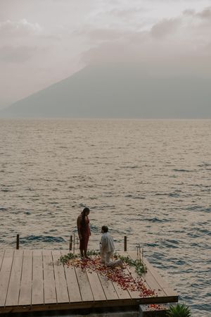Two figures stand on a wooden dock scattered with rose petals overlooking a misty lake and mountain landscape at dusk.