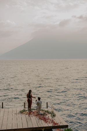 A romantic lakeside dock decorated with rose petals against a misty mountain backdrop at sunset.