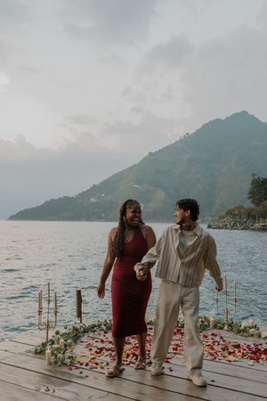 A romantic couple stands on a flower-covered dock overlooking a misty lake and mountain landscape at dusk.