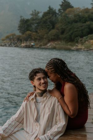 Intimate couple sharing tender moments on a wooden dock by a serene lake with trees in the background.