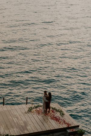 A sequence of photos showing a couple embracing on a wooden dock by the ocean with scattered red rose petals.