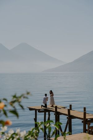 A wooden dock extends into a serene lake with misty mountains in the background and flowers in the foreground.