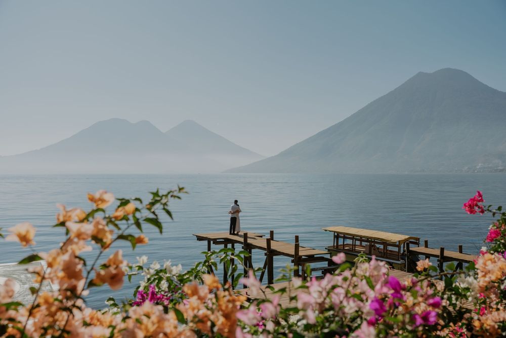 Scenic view of Lake Atitlan in Guatemala with wooden dock, pink bougainvillea flowers and volcanic mountains in background.