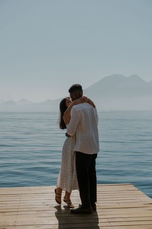 A couple embraces on a wooden dock overlooking misty mountains and calm waters at dawn.