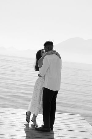 A romantic black and white scene of two people embracing on a seaside pier.