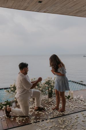 Man proposes on one knee to woman on oceanfront deck adorned with white flower petals and candles at dusk.