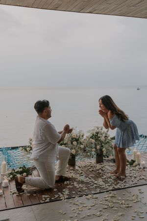 Man proposes to woman on one knee by the ocean, surrounded by white flowers, rose petals, and candles at dusk.