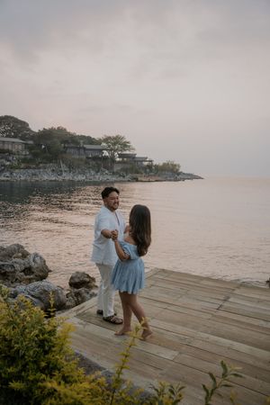 Couple holding hands on a wooden dock by the ocean at sunset, with rocky shoreline and houses in the background.