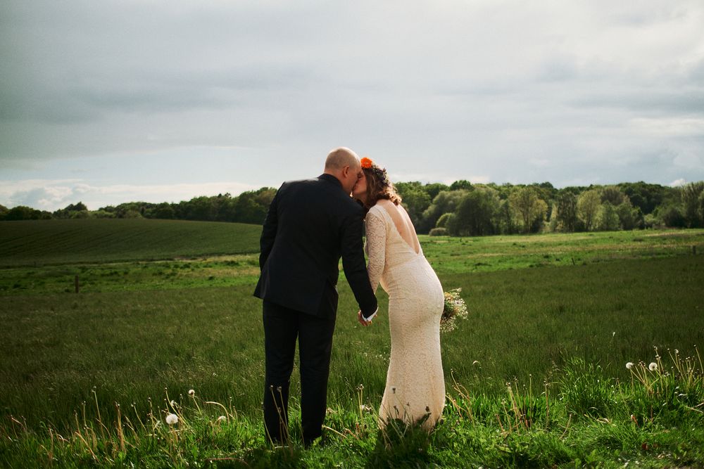 A couple in formal wedding attire stands together in a lush green meadow under cloudy skies.