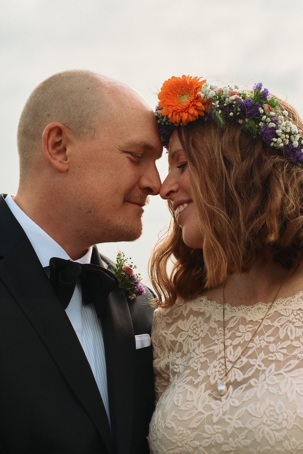 A romantic wedding moment captured with the bride wearing a floral crown and lace dress next to groom in black tuxedo.