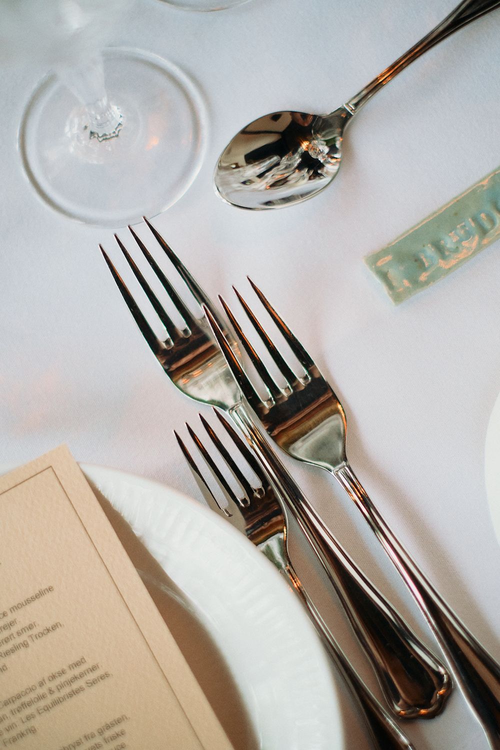 Elegant place setting with polished silver forks, spoon and menu card on crisp white tablecloth at upscale restaurant.