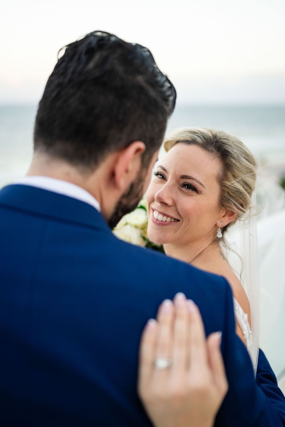 A radiant bride in a white veil smiles lovingly at her groom in a navy suit during their seaside wedding.