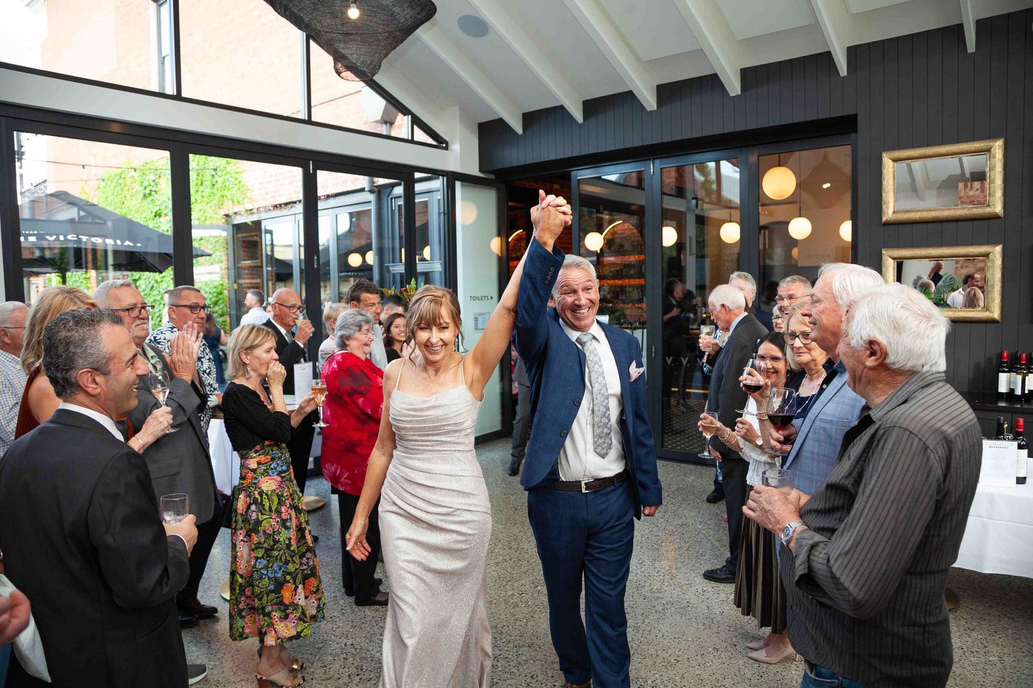 Wedding guests mingle and socialize during an indoor reception with string lights and wooden beams overhead.