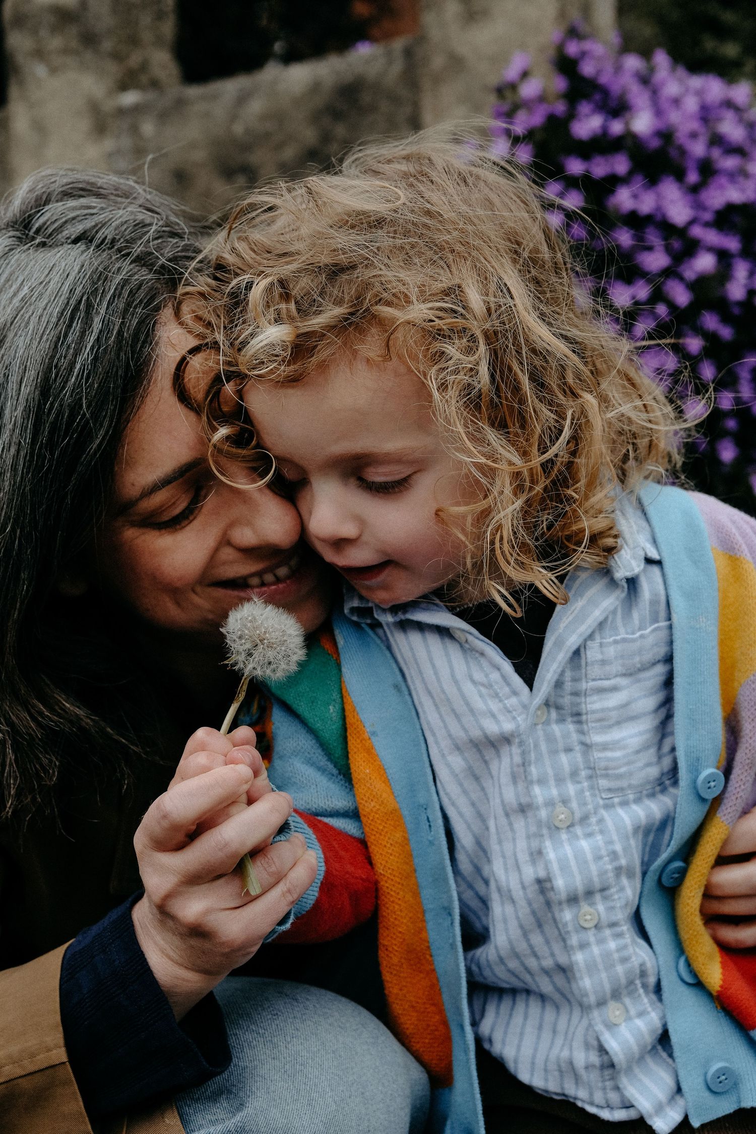 A mother and curly-haired child sharing a joyful, candid moment outdoors, with purple flowers softly in the background.