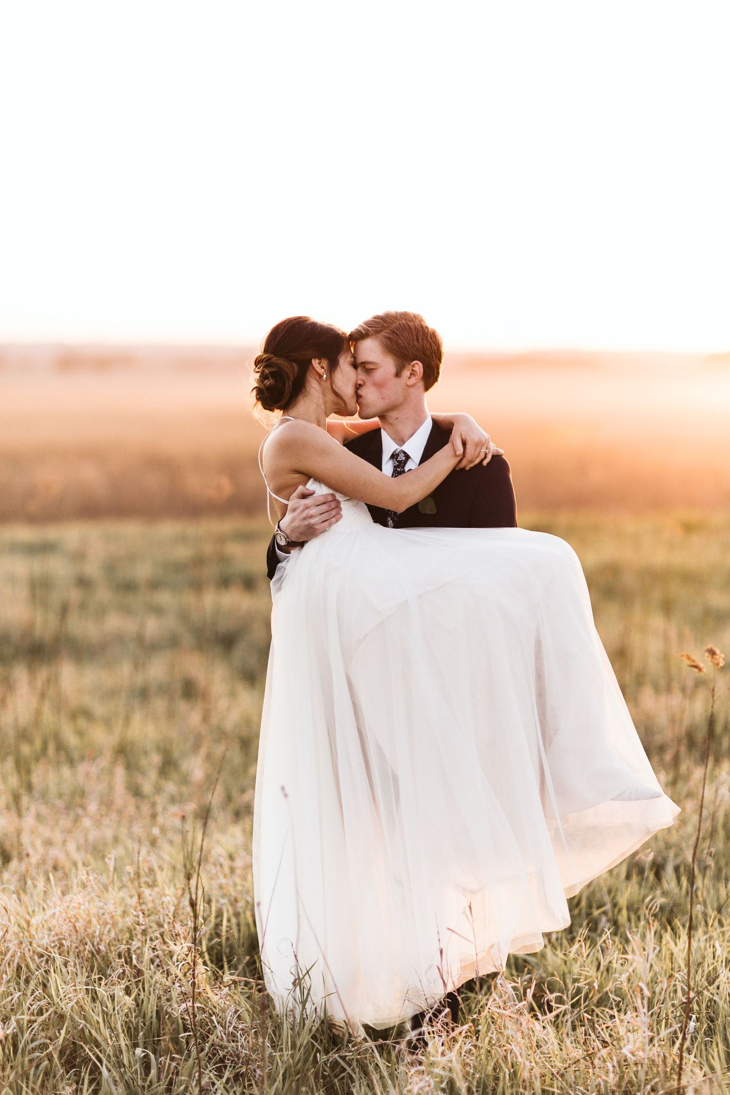 A romantic couple embraces in a golden field at sunset, with the bride's white dress flowing in the breeze.