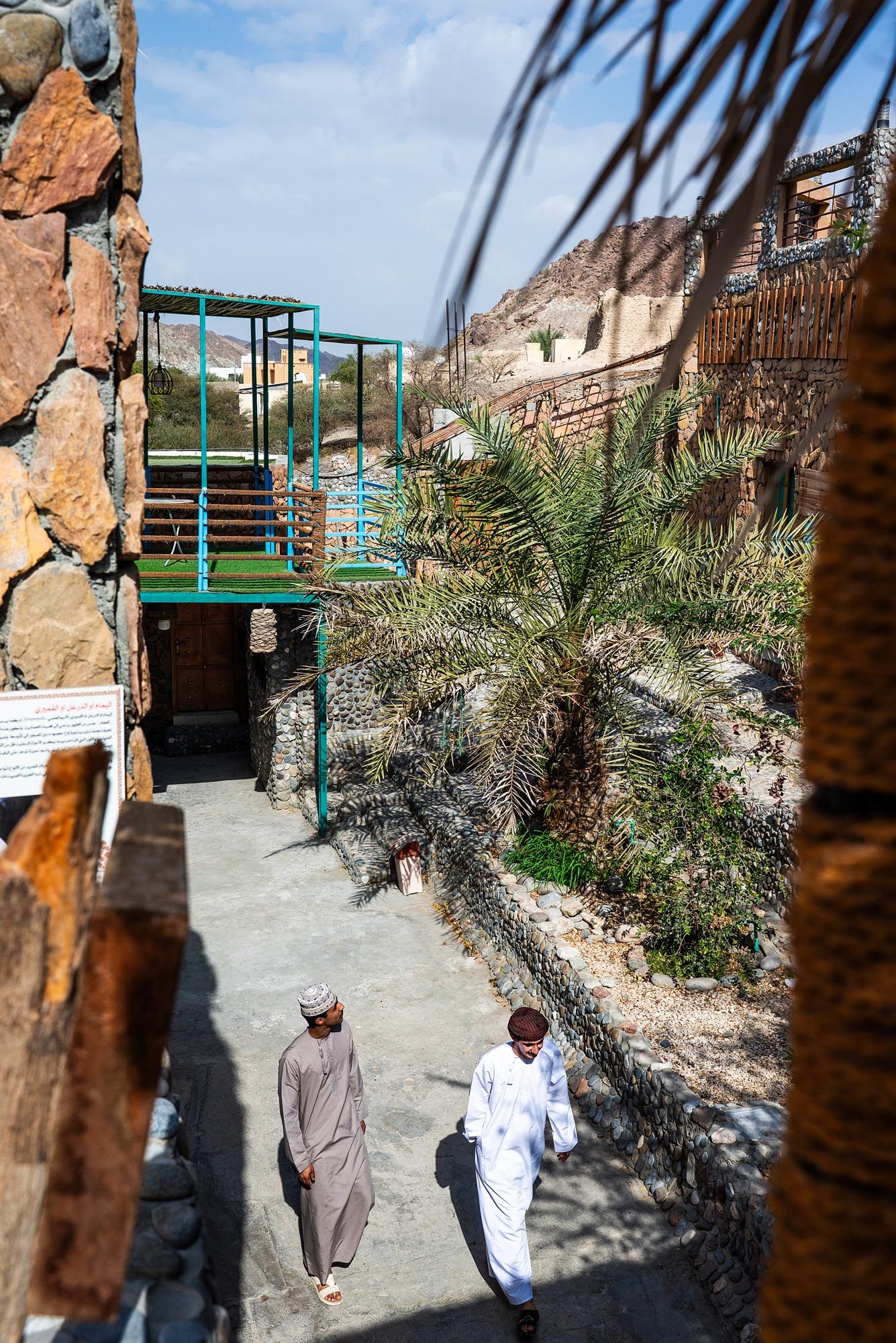 Visitors walk along a palm-lined pathway at a desert tourist attraction with rocky cliffs and green railings.