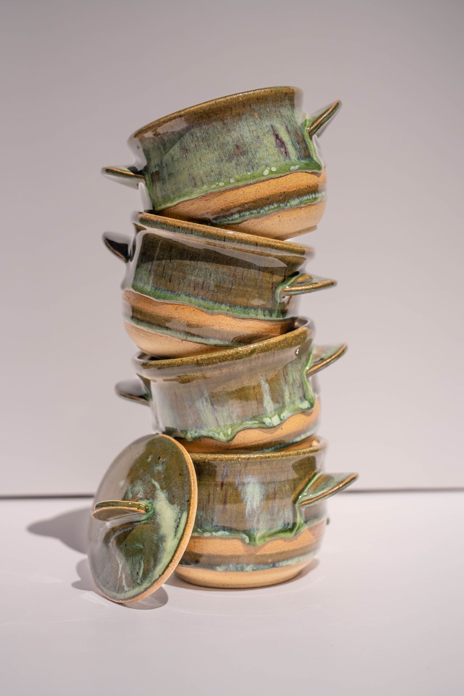 Stack of six handmade ceramic bowls with green and tan glaze, featuring small handles, balanced on a white surface.