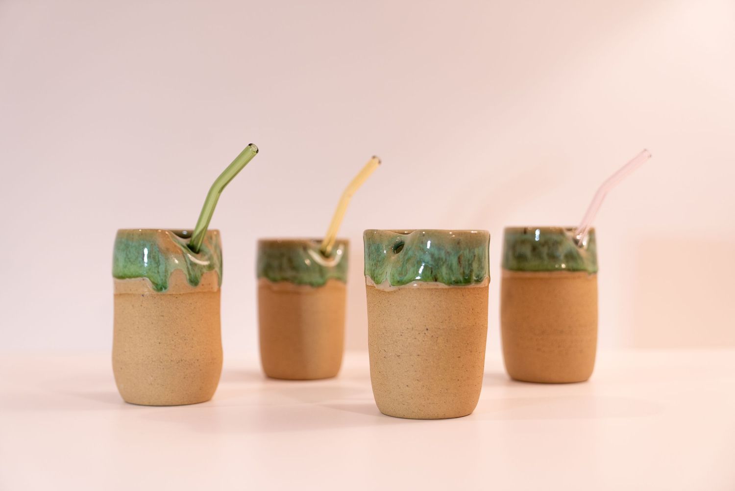 Four handmade ceramic cups with green drip glaze and colorful glass straws on a white background.