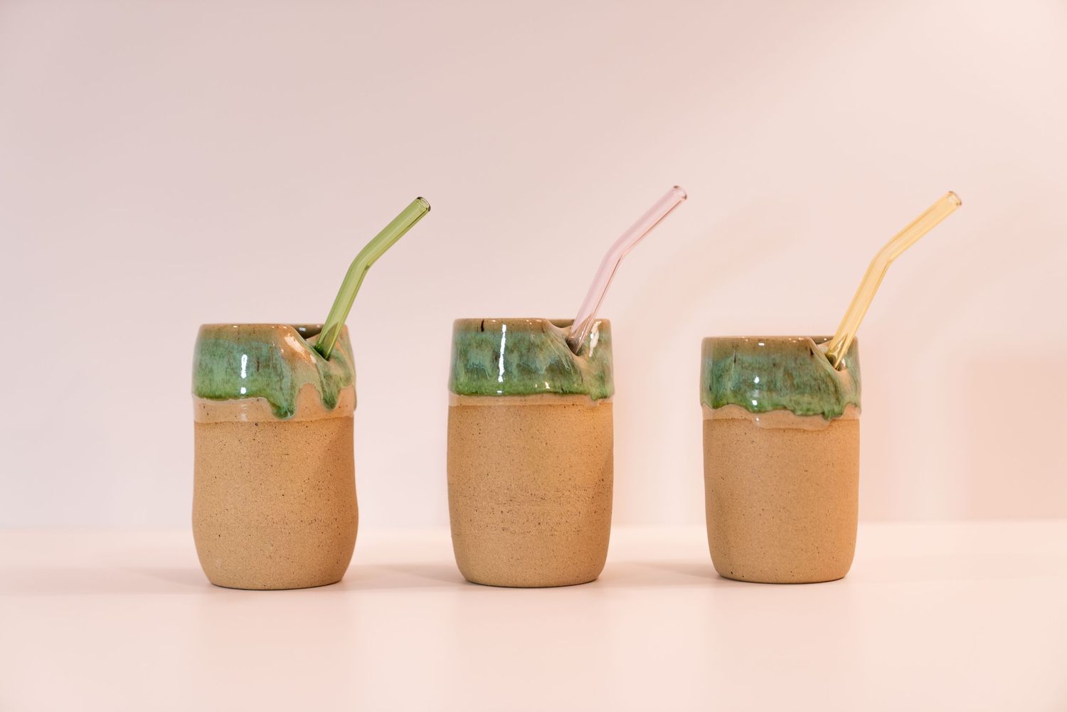 Three handmade ceramic cups with green drip glaze and colorful glass straws on a white background.