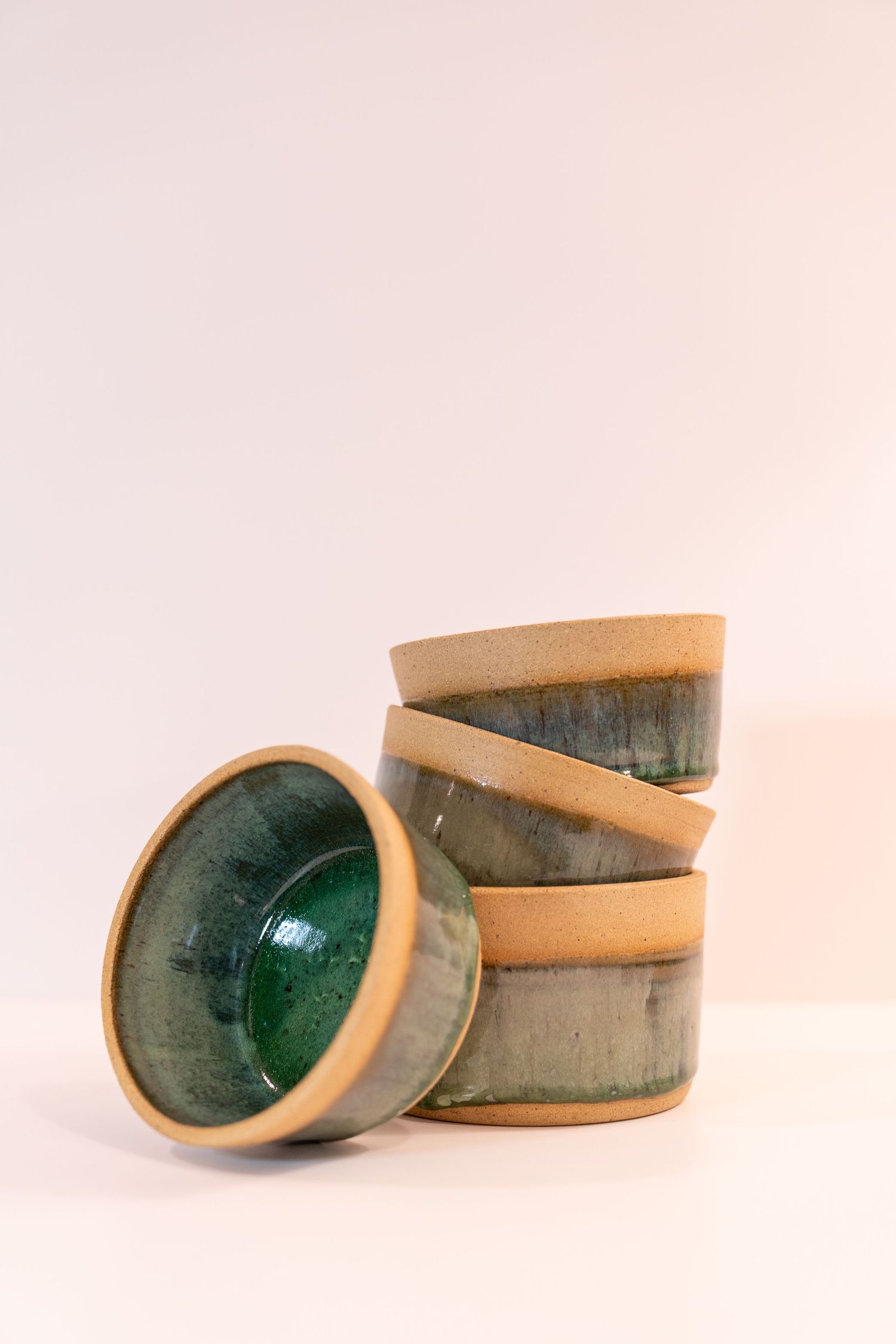 Stack of three handmade ceramic bowls with green glaze and natural clay rim, one leaning against stack on white background.