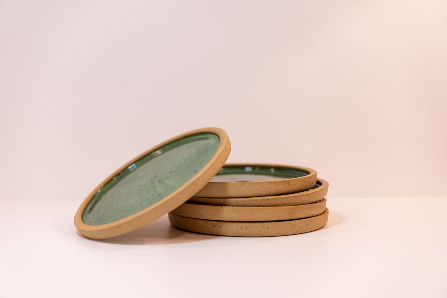 Stacked handmade ceramic plates with green speckled glaze and unglazed sandy rim with one leaning against the stack with a white background.